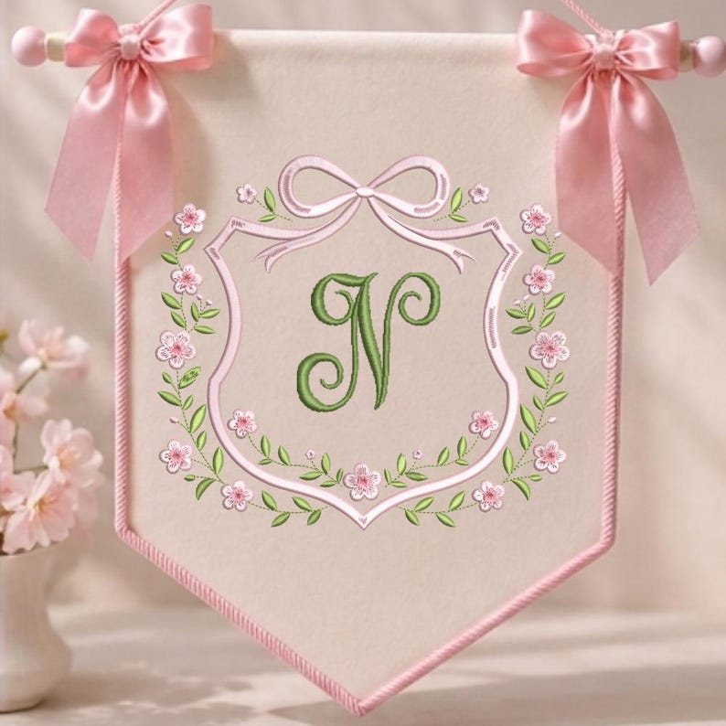 May include: A cream-colored fabric wall banner featuring a central embroidered green letter N. The banner is framed by a pink embroidered crest and floral wreath, finished with pink satin ribbon bows at the top corners on a wooden rod.