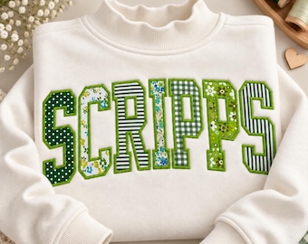 May include: A cream-colored sweatshirt featuring the word SCRIPPS in large, block-style letters. Each letter is filled with a different green and white pattern, including polka dots, floral prints, and stripes, all outlined in green thread.