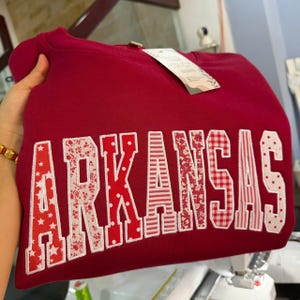 May include: A maroon crewneck sweatshirt featuring the word ARKANSAS in large, block-style applique letters. Each letter is crafted from different red and white patterned fabrics, including stars, floral prints, polka dots, and gingham checks.