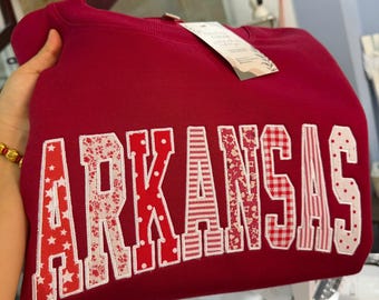 May include: A maroon crewneck sweatshirt featuring the word ARKANSAS in large, block-style applique letters. Each letter is crafted from different red and white patterned fabrics, including stars, floral prints, polka dots, and gingham checks.