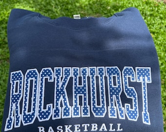 May include: A navy blue crewneck sweatshirt featuring the word ROCKHURST in large, white-outlined block letters with a blue and white polka dot pattern. Below the main text, the word BASKETBALL is printed in a smaller, simple white font.