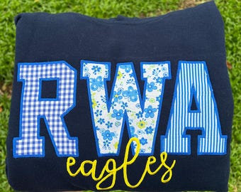 May include: A navy blue sweatshirt featuring large block letters RWA appliqued in different patterns: a blue and white gingham R, a blue and white floral W, and a blue and white striped A. Below, the word eagles is embroidered in yellow script.
