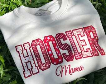 May include: A white crewneck sweatshirt featuring the word HOOSIER in large, block-style letters with various red and white patterns, including polka dots, stars, and floral prints. Below the word, the text Mama is embroidered in red script.
