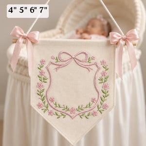 May include: A cream-colored fabric wall banner featuring an embroidered pink shield outline topped with a bow and surrounded by green vines and pink flowers. The banner hangs from a wooden rod with pink satin bows and includes the text 4" 5" 6" 7".