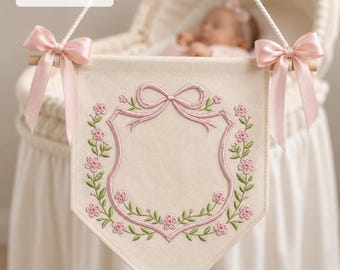 May include: A cream-colored fabric wall banner featuring an embroidered pink shield outline topped with a bow and surrounded by green vines and pink flowers. The banner hangs from a wooden rod with pink satin bows and includes the text 4" 5" 6" 7".