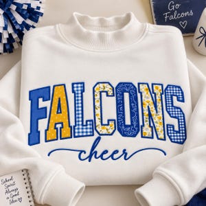 May include: A white long-sleeve sweatshirt with a mock neck collar. The front features the word FALCONS in large block letters with varied yellow, blue, and floral patterns, and the word cheer in blue cursive script underneath.