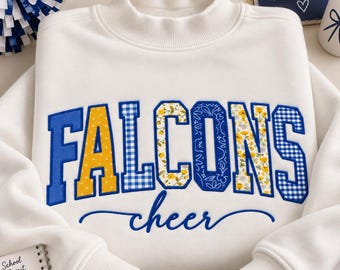 May include: A white long-sleeve sweatshirt with a mock neck collar. The front features the word FALCONS in large block letters with varied yellow, blue, and floral patterns, and the word cheer in blue cursive script underneath.