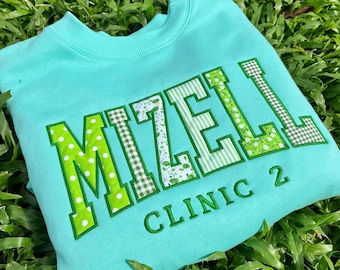 May include: A light turquoise crewneck sweatshirt featuring the name MIZELL in large, varsity-style applique letters. Each letter is filled with varying green and white patterns, including polka dots, gingham, and floral prints. Below, CLINIC 2 is embroidered in green.