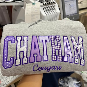 May include: A heather gray hooded sweatshirt featuring the word CHATHAM in large, purple-outlined block letters with varied purple and white patterns, including polka dots, florals, gingham, and stripes, with the word Cougars embroidered below.