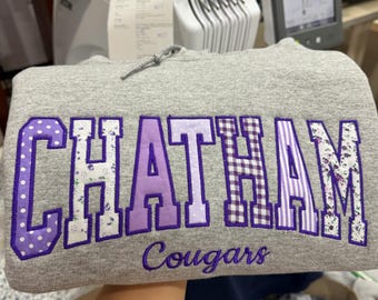 May include: A heather gray hooded sweatshirt featuring the word CHATHAM in large, purple-outlined block letters with varied purple and white patterns, including polka dots, florals, gingham, and stripes, with the word Cougars embroidered below.