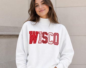 May include: A white mock neck sweatshirt featuring the word WISCO in large, red, block-style letters. Each letter has a distinct pattern, including white polka dots, small floral prints, and a red and white gingham check design on the final letter.