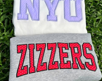May include: A white sweatshirt featuring the letters NYU in light purple with white polka dots and a grey sweatshirt featuring the word ZIZZERS in red with white polka dots, both with stitched outlines on the fabric surface.