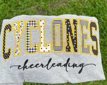 May include: A heather gray sweatshirt featuring the word CYCLONES in large, block-style letters with varied black and white patterns, outlined in yellow. Below, the word cheerleading is embroidered in a black, cursive script font.