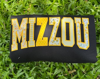 May include: A black sweatshirt featuring the word MIZZOU in large block letters. Each letter is outlined in yellow and filled with a variety of patterns, including yellow polka dots, light blue stripes, and small yellow floral prints.