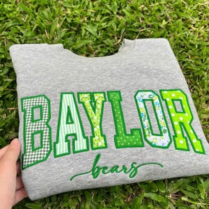 May include: A heather gray crewneck sweatshirt featuring the word BAYLOR in large, block-style letters. Each letter has a unique green and white pattern, including gingham, stripes, floral, and polka dots. The word bears is written below in cursive.