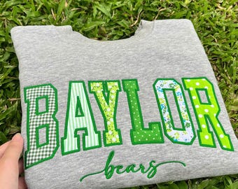 May include: A heather gray crewneck sweatshirt featuring the word BAYLOR in large, block-style letters, each with a unique green and white pattern, and the word bears written below in a green cursive script font.