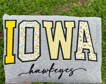 May include: A heather gray sweatshirt featuring the word IOWA in large block letters with black outlines. The letters I, O, and A have a yellow and white floral pattern, while the letter W has a yellow and white gingham pattern. The word hawkeyes is embroidered in black cursive script below.