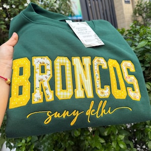 May include: A dark green crewneck sweatshirt featuring the word BRONCOS in large, yellow-bordered block letters with alternating floral and gingham patterns. Below, the text suny delhi is embroidered in yellow cursive script. A white tag is attached.