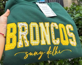 May include: A dark green crewneck sweatshirt featuring the word BRONCOS in large, yellow-bordered block letters with alternating floral and gingham patterns. Below, the text suny delhi is embroidered in yellow cursive script. A white tag is attached.