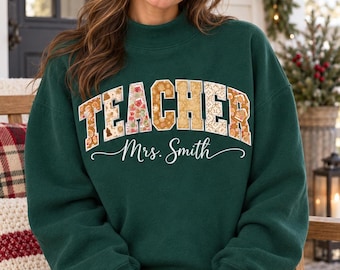 May include: A dark green long-sleeve sweatshirt with a mock neck. The word TEACHER is printed in a large, arched, patterned font across the chest, with the name Mrs. Smith written in white cursive script underneath the text.