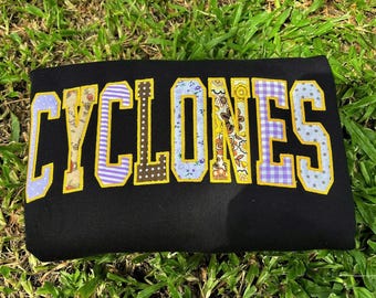 May include: A black sweatshirt featuring the word CYCLONES in large, block-style letters. Each letter is filled with a different patterned fabric, including floral, polka dot, and gingham prints, and is outlined in a thick, bright yellow thread.