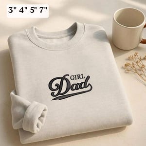 May include: A beige crewneck sweatshirt featuring black embroidered text that reads "Girl Dad" in a stylized script font. The image includes a text overlay indicating available sizes of 3", 4", 5", and 7" for the embroidery design.