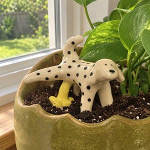 May include: A ceramic dog figurine with a cream-colored body and black polka dots, depicted in a lifting-leg pose. A small, bright yellow ceramic stream extends from the dog toward the soil, creating a humorous garden ornament for a plant pot.
