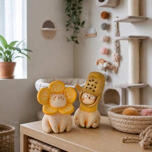May include: Two small, light-colored cat figurines. One cat wears a large, yellow, five-petaled flower collar around its face. The other cat wears a yellow, perforated, shoe-shaped head covering that leaves its face visible.