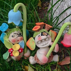 Könnte beinhalten: Zwei kleine, blasse, tonartige Figuren mit großen Augen und strukturiertem grünem Haar. Eine hält eine blaue glockenförmige Blume mit gelben Akzenten. Die andere trägt orangefarbene pilzartige Kopfbedeckungen und hält eine rosa Glockenblume.