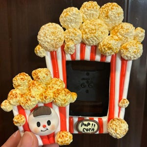 May include: A rectangular picture frame decorated with sculpted popcorn and red and white vertical stripes. A smaller popcorn bucket attachment features a cartoon face and the text Pop!! corn on a small oval plaque at the bottom of the frame.