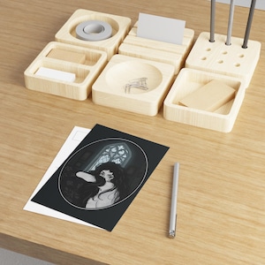 May include: A light-colored wooden desk organizer set with multiple compartments containing a roll of tape, paper clips, and pens, alongside a rectangular postcard featuring a black and white illustration of a person before a gothic window.