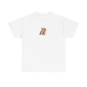 Może przedstawiać: Biały t-shirt z krótkim rękawem z małą, wyśrodkowaną grafiką przedstawiającą brązową kapibarę w szaliku w pionowe niebieskie, białe i czerwone paski. Koszulka ma okrągły dekolt i standardowy krój, prezentując prosty i przejrzysty design.