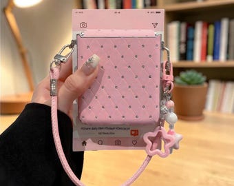 Puede incluir: Una funda de teléfono rectangular de color rosa claro con un patrón de diamantes acolchado y pedrería incrustada. La funda cuenta con una correa de muñeca rosa a juego y un dije colgante en forma de estrella. Está montada sobre una tarjeta con interfaz social.