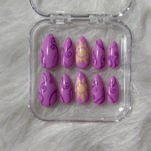 May include: A set of ten almond-shaped press-on nails in a clear plastic storage case. The nails are light purple with darker purple swirling line patterns and dotted accents. Two of the nails feature a central yellow sun design with rays.