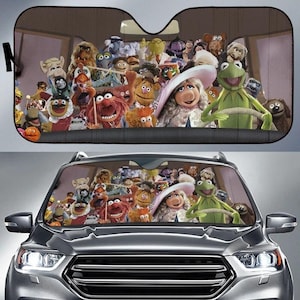 May include: A car windscreen sun shade featuring a colourful, crowded graphic print of various Muppet characters, including Kermit the Frog and Miss Piggy, positioned as if they are sitting inside the vehicle looking out through the front window.