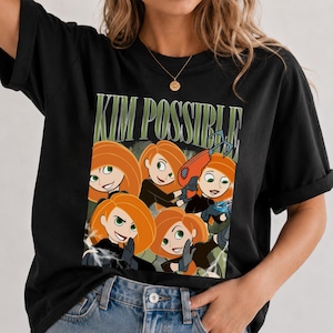 以下が含まれることがあります： 様々なポーズをとるキャラクター「キム・ポッシブル」のグラフィックプリントが施された黒の半袖Tシャツ。キャラクターのイラストの上には、大きな緑色の文字で「KIM POSSIBLE」とプリントされており、グラップリングフックのツールも描かれています。