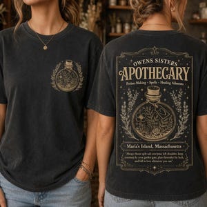 Puede incluir: Una camiseta gris carbón con un pequeño gráfico en el pecho izquierdo y un gran diseño rectangular en la espalda. El estampado trasero muestra el texto "OWENS SISTERS APOTHECARY" con una ilustración de una botella y detalles botánicos.