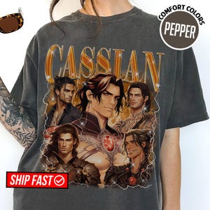 Op de afbeelding: Een antracietgrijs T-shirt met korte mouwen en een grafische print van vijf mannelijke personages met lang donker haar en de tekst CASSIAN in grote gouden letters. Het ontwerp bevat bliksemschichten en een cirkelvormig embleem met de tekst COMFORT COLORS PEPPER.