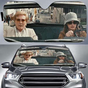 May include: A car windshield sunshade featuring a printed scene of two people in a convertible car. One person wears a beige blazer and glasses, while the other wears a grey bucket hat and sunglasses. The scene depicts a city street background.
