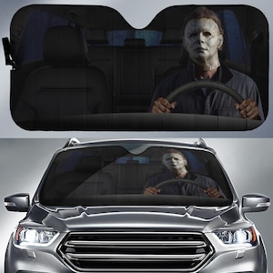May include: A car windscreen sunshade featuring a graphic print of a person wearing a white mask and a dark shirt, appearing to sit in the driver seat and hold a steering wheel. The design creates an illusion of a passenger inside the vehicle.