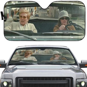 May include: A car windshield sunshade featuring a printed scene of two people in a convertible car. The person on the left wears a beige blazer and glasses, while the person on the right wears a brown jacket, a grey bucket hat, and sunglasses.