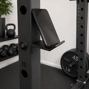 May include: A matte black L-shaped phone holder mount attached to a vertical steel power rack post. The holder features a flat base and a small front lip to secure a smartphone in an upright position. The rack post has circular holes and the number 15.