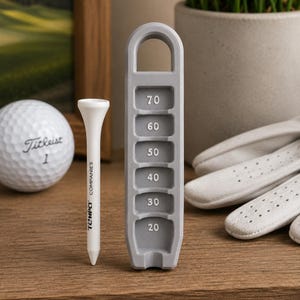 Puede incluir: Una pelota de golf blanca con el texto Titleist 1, un tee de golf blanco con el texto TEMPO COMPANIES y un soporte de plástico gris para tees con una columna vertical de ranuras numeradas del 20 al 70, en un diseño lineal y funcional.