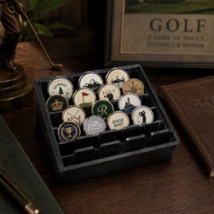 May include: A black compartmentalized display tray containing fourteen circular golf ball markers. Each marker features distinct designs including a trophy, a lighthouse, a compass, a mountain, a golf flag, and text such as Eagle and Good Shot.