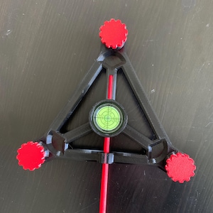 Puede incluir: Una herramienta de nivelación triangular negra con tres perillas de ajuste rojas en cada esquina y un nivel de burbuja circular central con líquido verde. Una varilla roja delgada se extiende desde el centro del marco, proporcionando una referencia visual clara para tareas de alineación y nivelación de precisión.