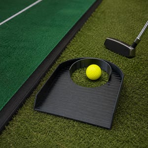 May include: A bright yellow golf ball rests inside a black plastic putting cup training aid. The cup features a flat base with a circular opening and a raised back wall. A silver golf putter head is positioned nearby on the green turf surface.