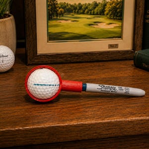 May include: A white golf ball held within a red circular plastic frame attached to a grey Sharpie fine point permanent marker. The golf ball features a thin blue horizontal line drawn across its center, and the marker displays black text.