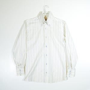 Puede incluir: Una camisa blanca de manga larga con botones y finas rayas azules verticales. La camisa tiene un cuello clásico y puños con botones.