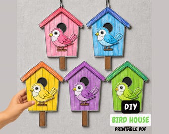 Op de afbeelding: Vijf hangende vogelhuisdecoraties in roze, blauw, geel, paars en groen, elk met een bijpassende gekleurde vogel op een bruine houten pin. De tekst DIY BIRD HOUSE PRINTABLE PDF staat in de rechterbenedenhoek.