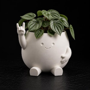 May include: A matte white ceramic planter shaped like a round character with a smiling face, two small feet, and two arms. One arm is raised in a rock-and-roll hand gesture, while the other rests on its side. A green Peperomia plant grows inside.